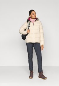 Beige Steppjacke mit Kapuze, lila Innenfutter, über einem pinkfarbenen Oberteil getragen. Kombiniert mit dunkler Hose und braunen Wanderschuhen. Schwarzer Rucksack.