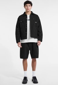 Chaqueta de mezclilla negra con dos bolsillos en el pecho, combinada con una camiseta blanca y pantalones cortos de mezclilla negra. Unos zapatos negros y calcetines blancos completan el look.
