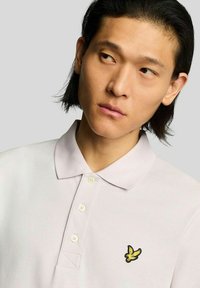 Lichtpaarse poloshirt met een kraag, met een geel geborduurd logo van een vogel op de linkerborst. Zachte stof en knoopdetails.