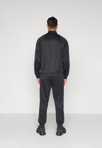 Μαύρη φόρμα με μοβ ρίγες στα πλάγια, κατασκευασμένη από λεία ύφασμα· διαθέτει ζακέτα με φερμουάρ και ελαστικές μανσέτες και κάτω μέρος στα παντελόνια.