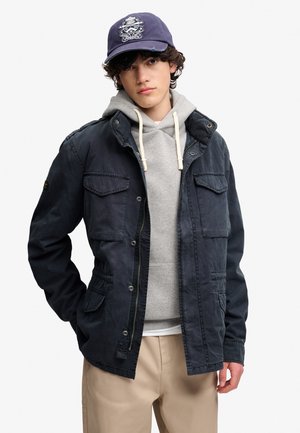 Ung mand iført navyblå kasket, sort jakke over grå hættetrøje og beige bukser står med den ene hånd i lommen.