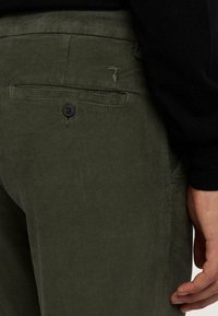 Olivgrön kordfläskbyxor med vertikal ribbning, med en bakficka som har en svart knapp och en liten logotyp ovanför fickan.