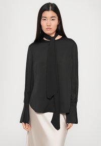 Črna bluzica z dolgimi rokavi, izdelana iz lahkotnega materiala, z zavezovanjem za šal okoli vratu, širokimi manšetami in ukrivljenim robom, kombinirana s svetlo satensko krilom.