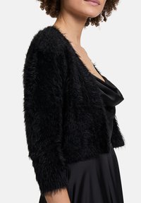 Schwarze, kurz geschnittene Strickjacke aus flauschigem Material. Verfügt über Dreiviertelärmel und eine weiche Textur, getragen über einem schwarzen Satin-Kleid.