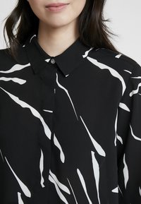Schwarze Bluse mit einem geknöpften Kragen, verziert mit weißen abstrakten Streifenmustern. Glattes, leichtes Material mit lässiger Passform.