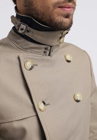 Gros plan sur un homme portant un manteau beige croisé avec de gros boutons et un col intérieur noir fixé par une boucle.