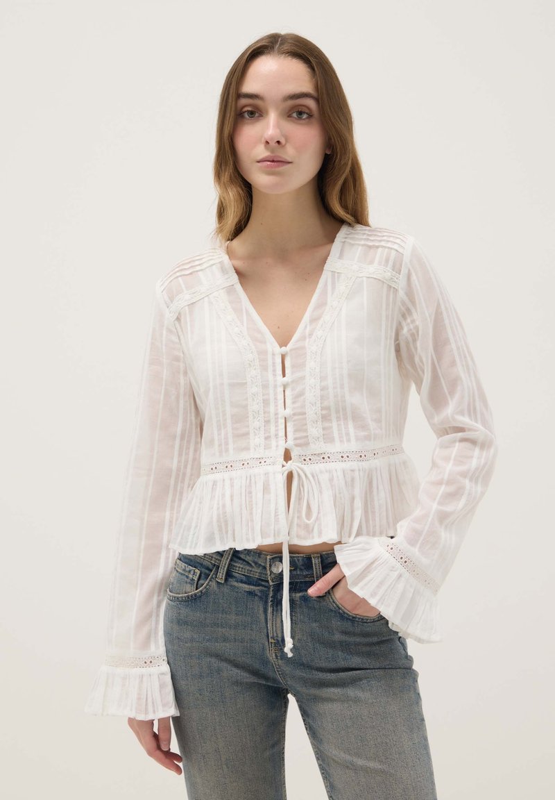 Joven mujer vistiendo una blusa blanca transparente con detalles de encaje, puños con volantes y un lazo en la cintura, combinada con pantalones vaqueros azules.