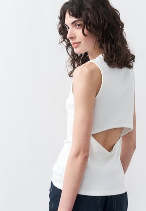 Ženska z kodrastimi lasmi, oblečena v belo majico brez rokavov z trikotno odprtino na hrbtu, kombinirano s temnimi hlačami, na belem ozadju.