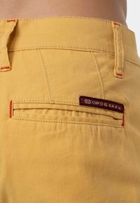 I pantaloni gialli presentano cuciture rosse a contrasto e una tasca posteriore con un'etichetta del marchio color bordeaux. Il tessuto appare liscio e resistente.