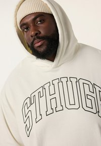 Cremfarbener Hoodie mit auffälligen schwarzen "STHUG"-Buchstaben. Das Material scheint weich und locker geschnitten zu sein. Begleitet von einer beigen Strickmütze.