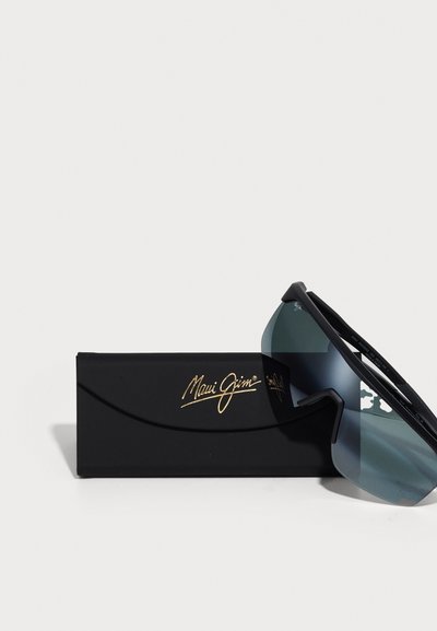 Solglasögon med en svart ram och grönaktiga linser, placerade på ett svart fodral med en guldfärgad "Maui Jim"-logotyp.