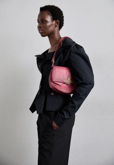 Saco de couro na cor baga com um formato curvo, ferragens douradas e uma alça. O modelo usa um blazer estruturado preto e calças.