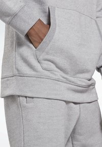 Main dans la poche d'un sweat à capuche gris clair texturé porté avec un pantalon assorti, montrant en gros plan les détails du tissu et les coutures.