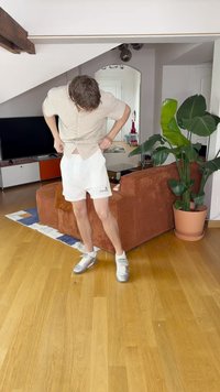 Biele šortky s čiernym logom, svetlošedá pruhovaná polokošeľa s krátkym rukávom a strieborné tenisky. Interiér s zelenou rastlinou.