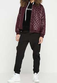 Bordeaux gevoerd bomberjack met geribde manchetten, gedragen over een zwarte hoodie, gecombineerd met zwarte tapered broek en witte sneakers.