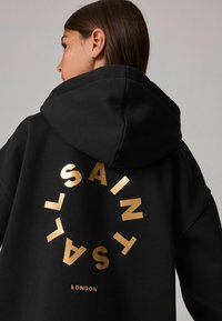 Sudadera negra con capucha y un logo circular dorado que presenta el texto "SAINT" y "LONDON" en la parte posterior. Tela suave, corte relajado y con capucha.