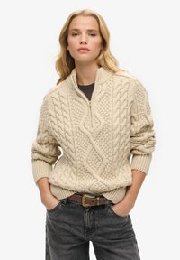 Beige kabelbrei trui met een hoge kraag, voorzien van textuurpatronen en geribbelde boorden, gecombineerd met donkere spijkerbroek.