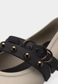 Chaussure plate crème avec une bride en cuir noir ornée de clous dorés et une fermeture à boucle dorée.
