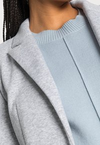 Blazer gris clair porté sur un haut en maille côtelée bleu clair. Le blazer présente des revers crantés ; le haut a une finition texturée avec un col rond.
