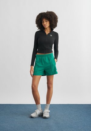 Νέα γυναίκα με σγουρά μαλλιά στέκεται σε μπλε χαλί φορώντας μαύρο μακρυμάνικο τοπ με φερμουάρ, πράσινο αθλητικό σορτσάκι, γκρι κάλτσες και γκρι αθλητικά παπούτσια.