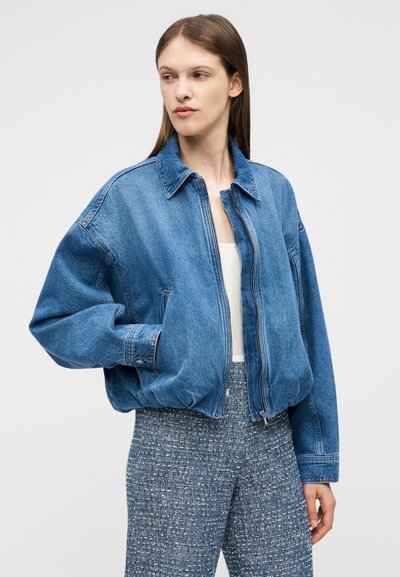 Femme portant une veste en denim bleue oversize par-dessus un haut blanc et un pantalon gris texturé, debout avec une main dans la poche, regardant vers la gauche.