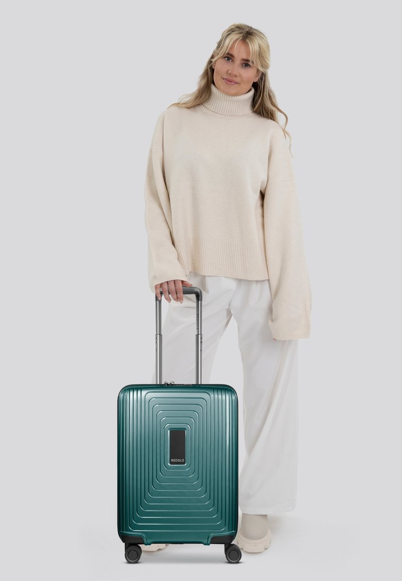 Valigia rigida color teal con design a nervature, maniglia telescopica e quattro ruote. Posizionata accanto a una persona in un maglione beige e pantaloni bianchi.
