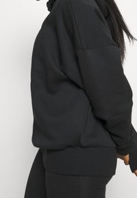 Hoodie noir surdimensionné avec une texture douce, présentant une poche kangourou et des poignets côtelés. La construction sans couture met en avant le confort.