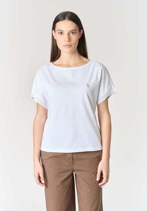 Donna con capelli lunghi e lisci, che indossa una maglietta bianca a maniche corte e pantaloni marroni, in piedi contro uno sfondo chiaro e semplice.