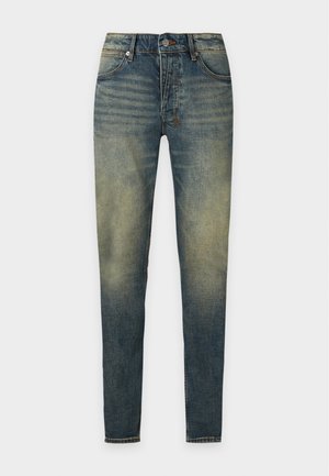 Jeune homme portant une veste noire à manches graphiques, un jean slim vert délavé et des mocassins noirs à pampilles, debout avec une main dans la poche.