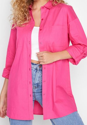 Frau trägt ein leuchtend pinkes, übergroßes Hemd über einem weißen Croptop und hoch taillierte blaue Jeans, mit einer Hand hält sie den vorderen Teil des Hemdes.