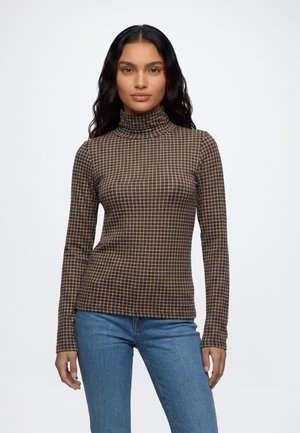 Frau mit langen dunklen Haaren, die einen braun-beigen karierten Rollkragenpullover und blaue Jeans trägt, steht vor einem schlichten weißen Hintergrund.