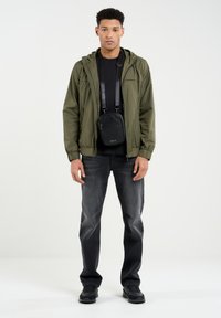 Alyvuogių žalia lengva striukė, juodas marškinėlis, tamsiai pilki džinsai ir juoda per petį nešama kuprinė. Modelis stovi prieš paprastą baltą foną.