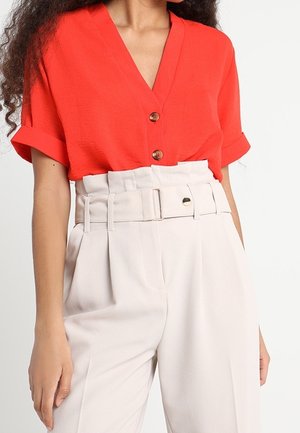 Femme portant un chemisier rouge vif à manches courtes avec un décolleté en V, des boutons marron et un pantalon blanc taille haute avec une large ceinture.