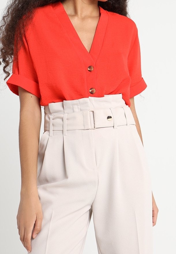 Femme portant un chemisier rouge vif à manches courtes avec un décolleté en V, des boutons marron et un pantalon blanc taille haute avec une large ceinture.