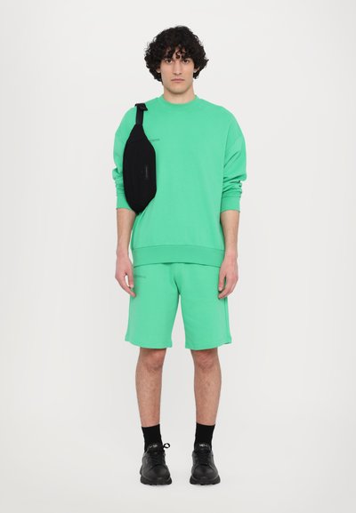 Sweatshirt en coton vert et shorts assortis, avec une coupe décontractée. Un sac crossbody noir est porté en bandoulière. Des sneakers noires complètent le look.