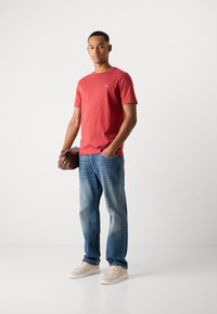 Camiseta de algodón roja de manga corta, jeans de mezclilla azul claro con acabado desgastado y zapatillas beige; sosteniendo un bolso de cuero marrón.