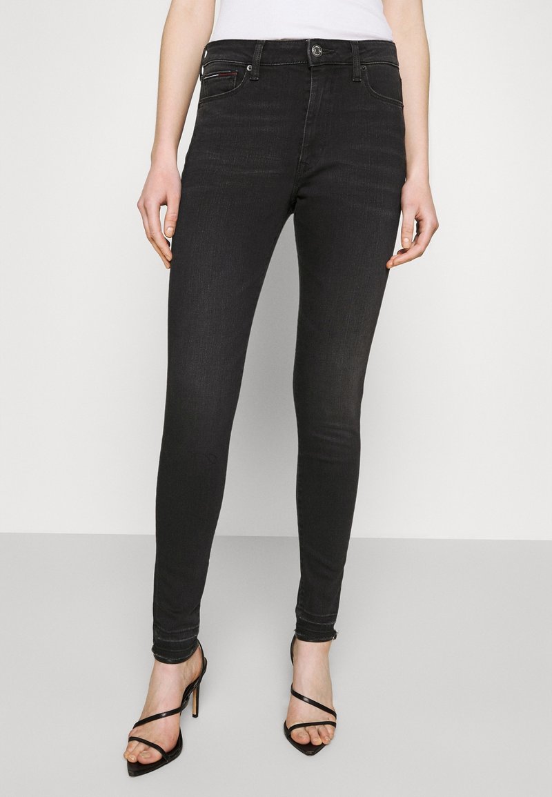 Jean skinny en denim noir avec taille mi-haute, doté d'une fermeture à bouton à l'avant, de cinq poches et d'une légère décoloration sur les jambes.