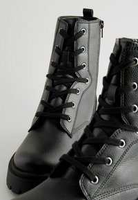Botas de cuero negro hasta el tobillo con un acabado texturizado, punta redonda, cordones frontales y cierre de cremallera lateral. Presentan ojales de metal y una suela gruesa.
