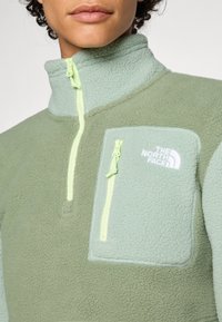 Groene fleecetop met een halve ritssluiting, voorzien van een klein borstzakje en contrasterende lichtgroene accenten.