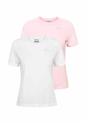 Mlada žena stoji, smiješi se, nosi bijelu majicu, tamnoplave traperice visokog struka, bijele čarape i bež tenisice, na jednobojnoj pozadini.