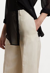 Chemise noire transparente à boutons superposée sur un pantalon large beige clair avec poches latérales, mettant en valeur une texture de tissu lisse et une coupe décontractée.