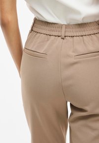 Beige byxor med resårmidja, med två bakfickor och en slät yta. Tyget hänger mjukt.