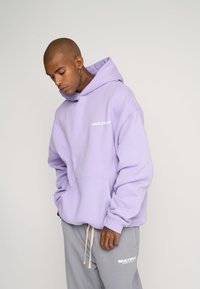 Sudadera de un suave tejido en color púrpura claro, con un bolsillo frontal y un logotipo blanco que dice "MULTIPLY". Combinada con pantalones de chándal grises que presentan el mismo logotipo.