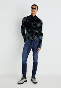 Svart og grønn strikket zip-up jakke, mørkeblå slim jeans og teksturerte grå sko. Modellen har på seg solbriller og står mot en lys bakgrunn.