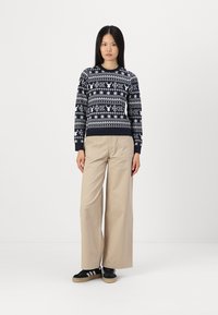 Pulover tricotat bleumarin cu modele de fulgi de zăpadă și reni albe, asortat cu pantaloni bej cu picioare largi și adidași negri cu dungi albe.