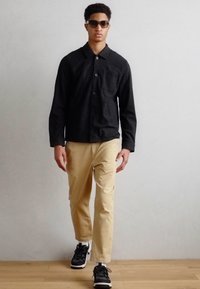 Casaco preto de botões, calças bege e ténis pretos usados por um modelo. Óculos de sol e um detalhe minimalista no bolso do casaco.