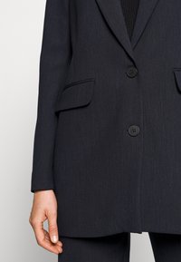 Mörkblå blazer i texturerad tyg, med slagkrage, två framfickor och två svarta knappar. Enkel design med rena linjer.
