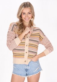 Cardigan lavorato a maglia in beige pastello con motivi geometrici multicolori, dotato di grandi bottoni e polsini a coste. Abbinato a pantaloni shorts in denim.
