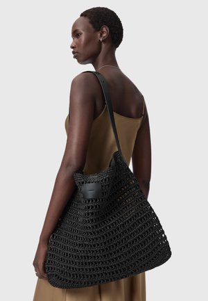 Γυναίκα με κοντά μαλλιά που φοράει ένα καφέ φόρεμα χωρίς μανίκια, κρατά μια μεγάλη μαύρη υφαντή τσάντα ώμου με δερμάτινο λουράκι.