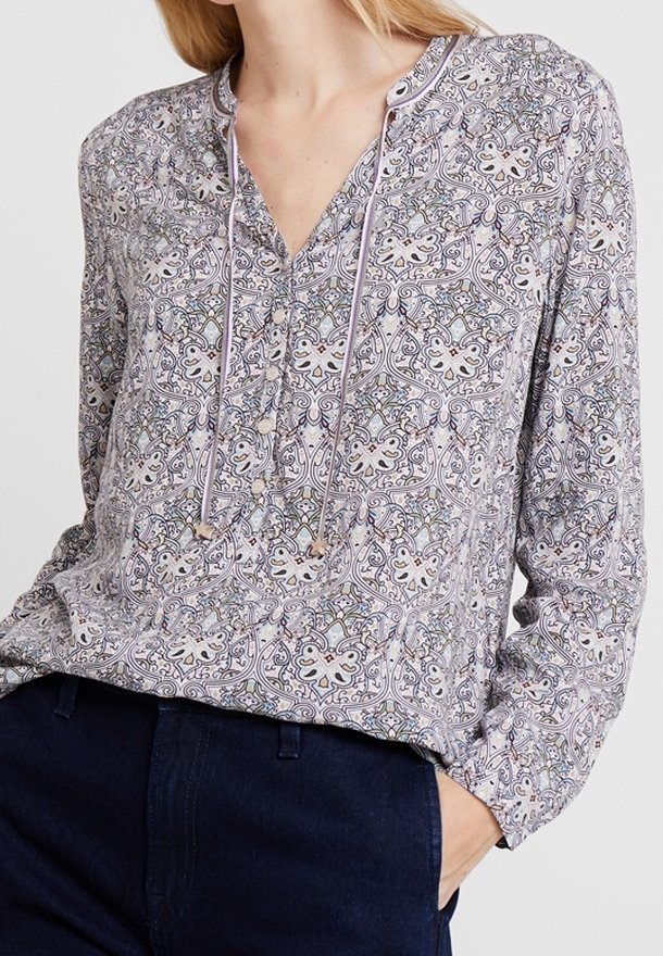 Bluzka z długim rękawem w jasnofioletowym kolorze, z misternymi wzorami kwiatowymi i geometrycznymi. Posiada dekolt w serek, detal ze sznureczkiem oraz miękką teksturę materiału.